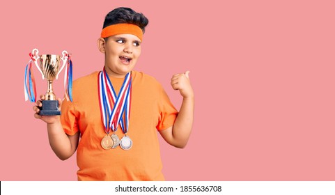 Little Boy Kid Holding Champion Trophy And Wearing Medals Pointing Thumb Up To The Side Smiling Happy With Open Mouth 