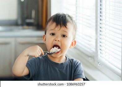 The Little Boy In The Kitchen Eagerly Eating Rice With A Spoon Independently