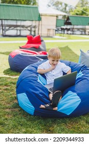 Little Boy With A Laptop On The Background Of Summer Grass. A Boy In A White T-shirt Is Sitting On A Beanbag In The Park.