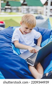 Little Boy With A Laptop On The Background Of Summer Grass. A Boy In A White T-shirt Is Sitting On A Beanbag In The Park.