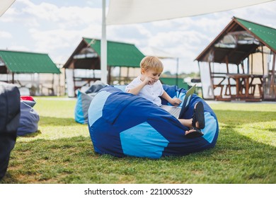 Little Boy With A Laptop On The Background Of Summer Grass. A Boy In A White T-shirt Is Sitting On A Beanbag In The Park.