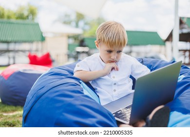 Little Boy With A Laptop On The Background Of Summer Grass. A Boy In A White T-shirt Is Sitting On A Beanbag In The Park.