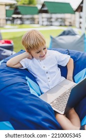 Little Boy With A Laptop On The Background Of Summer Grass. A Boy In A White T-shirt Is Sitting On A Beanbag In The Park.