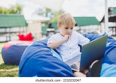 Little Boy With A Laptop On The Background Of Summer Grass. A Boy In A White T-shirt Is Sitting On A Beanbag In The Park.