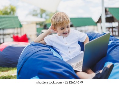 Little Boy With A Laptop On The Background Of Summer Grass. A Boy In A White T-shirt Is Sitting On A Beanbag In The Park.