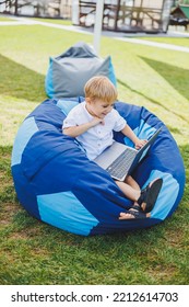 Little Boy With A Laptop On The Background Of Summer Grass. A Boy In A White T-shirt Is Sitting On A Beanbag In The Park.