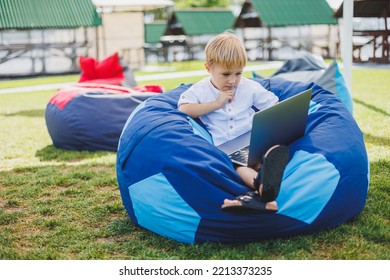 Little Boy With A Laptop On The Background Of Summer Grass. A Boy In A White T-shirt Is Sitting On A Beanbag In The Park.