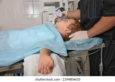 Little Boy Lies On The Operating Table Under A Ventilation Mask Held By A Doctor In Sterile Gloves. A Child Breathes Oxygen Through A Mask That Is In The Hands Of A Doctor