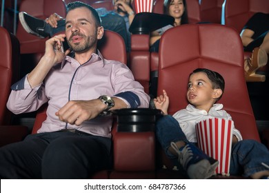 Little Boy Looking Disappointed With His Dad And Asking Him To Be Quiet While He Talks On The Phone At The Movie Theater