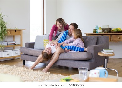 Little boy is lying in his father's lap on the sofa in the living room of their home. He is tickling him with help from the boys mother and his sister. 