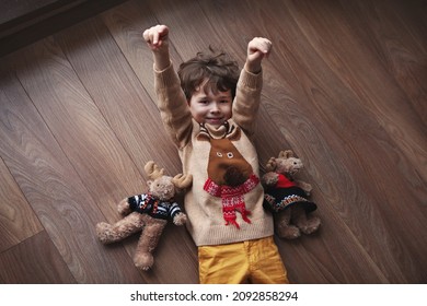 Little Boy is lying and smiling on wooden floor with plush toys, wearing sweater with reindeer - Powered by Shutterstock