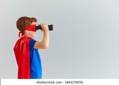 Little Boy In A Mask Cloak Of A Superhero Looks Through Binoculars Towards A Free Space For Text Standing In Profile On A Gray Background. Childrens Role Playing Games Concept.