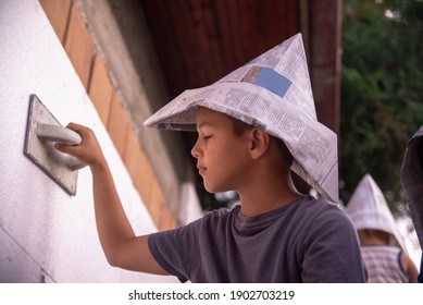 A Little Boy, A Master With A Newspaper Hat, Is Painting A House