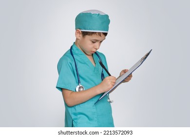 Little Boy In Medical Uniform Character Holds Clipboard And Writing Diagnosis. Serious Swarthy Boy Writes Down A Treatment Plan On Clip-on Tablet, Isolated On White