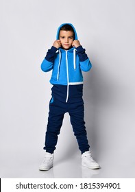 Little Boy Model Wearing Blue Black Sports Tracksuit And Pants Taking Off Hood. Full Length Isolated Studio Portrait On Gray Background. Male Child Sport Fashion Promotion