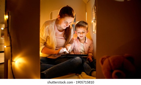 Little boy with mother playing in toy cardboard house and using tablet computer. Concept of child education and family having time together at night.