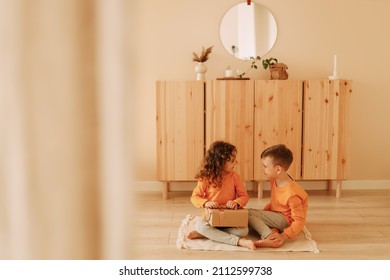 A little boy in orange pajamas congratulates and gives a gift box to a little girl in a cozy room with wooden furniture at home. Children friends celebrate birthday. Selective focus