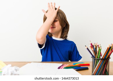 Little boy painting and doing homeworks on his desk forgetting something, slapping forehead with palm and closing eyes.