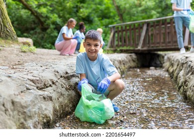 Little Boy Picks Up Some Recycling From The River. Family Collecting Recycling And Putting It In A Green Bag. Mother Teaching Kids About Sustainability.