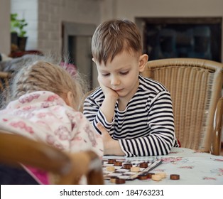 Little Boy Planning His Strategy Staring Thoughtfully At The Board As He Sits At A Table Playing A Game Of Checkers Or Draughts With His Sister