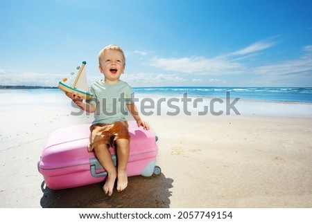 Little boy play hold sail boat yacht sitting on a suitcase on the beach vacations near the sea laughing