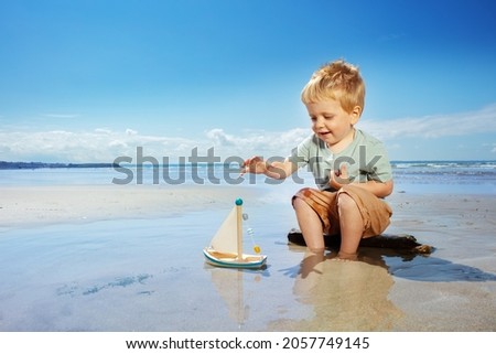 Little boy play with toy sail ship in the paddle sitting on the beach near sea