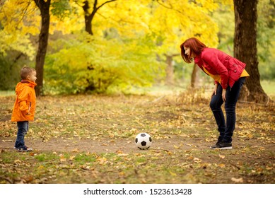 Little Boy Playing Football With Mother In Autumn Park. Mom Plays Soccer With Son Outdoors: Woman Leaned Forward Explaining Rules Of Game To Child, Ball Is Between Them. Yellow Trees In Background