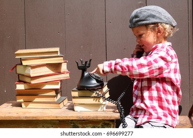 A Little Boy Is Playing Office, On An Old-fashioned Phone And A Stack Of Old Books. He Is Wearing A Hat Backwards And A Red And White Checkered Shirt. Labor Day, 