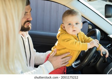 Little Boy Plays In The Car, Stands In Front Seat, Holding Wheel. Dad Supports Child. Father And Mom With Baby In The Car. Life Insurance, Parenthood, Happy Family, Trip, Small Driver Concept.