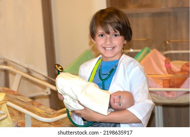 A Little Boy Plays Doctor With A Doll. Happy Boy Dressed As A Medical Officer Holds A Toy Baby In His Arms