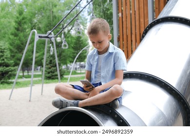 Little boy plays on his mobile phone instead of enjoying the park. Phone addiction. Child plays online chat on playground, against backdrop of nature, social problems, communication. - Powered by Shutterstock