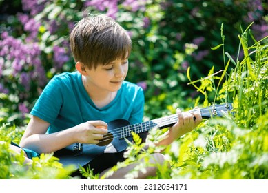 a little boy plays a small black ukulele, a guitar in a blooming garden, a park on a sunny day - Shutterstock ID 2312054171