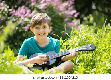 a little boy plays a small black ukulele, a guitar in a blooming garden, a park on a sunny day - Shutterstock ID 2312054213