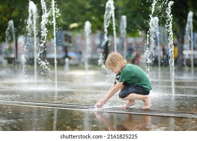 Little Boy Plays In The Square Between The Water Jets In The City Fountain At Sunny Summer Day. Active Summer Leisure For Kids In A Big City.