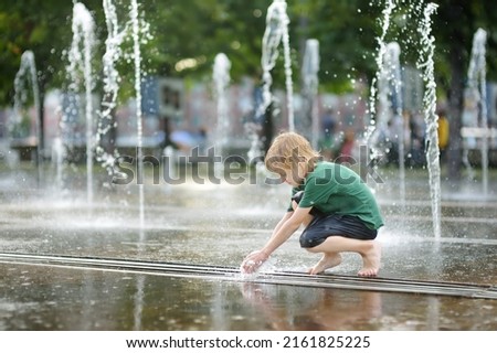 Little boy plays in the square between the water jets in the city fountain at sunny summer day. Active summer leisure for kids in a big city.