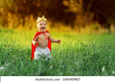 Little Boy Prince,child King Laughing Summer Day Outdoors Around Colorful Green Grass.Funny Knight Baby 1-2 Years.Cute Kid Smile Dressed Image Playing Hero,knight,warrior,red Cloak,gold Crown.