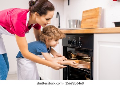 Little Boy Putting Tray Into Open Oven And His Mom Helping Him After Making Homemade Cookies On Weekend