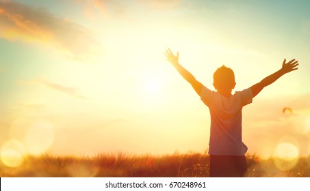 Little boy raising hands over sunset sky, enjoying life and nature. Happy Kid on summer field looking on sun. Silhouette of male child in sunlight rays. Fresh air, environment concept. Dream of flying