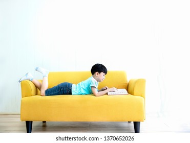 little boy read a book while laying down on sofa at home.Happy little boy reading red book while lying on his stomach on fabric yellow sofa.Portrait of Asian schoolboy read a book.