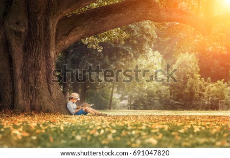 Little boy reading a book under big linden tree