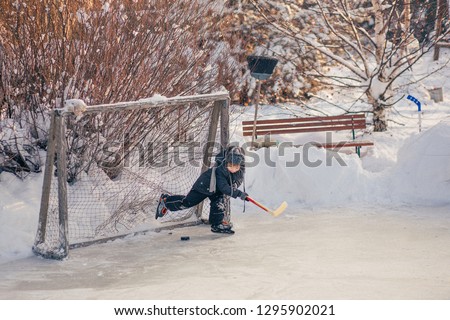 Little boy in retro clothes is playing hockey as a goalkeeper on the handmade rink in Russia village Image with selective focus, toning and noise effect.