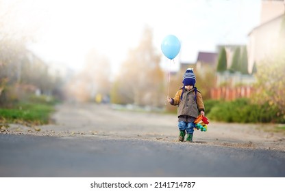 A little boy in a retro coat in a hat with a blue gel ball walks along the road. A cute boy warmly dressed with a balloon and a toy car is walking down the road. - Shutterstock ID 2141714787