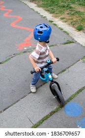 Little Boy Rides A Training Bike With His Back To The Camera In A Striped T-shirt, Wearing A Bike Helmet, In Front Of A Painted Red Winding Line.