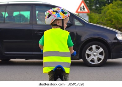 Little boy riding a bike on the road. Child is wearing reflective vest and helmet because of safety. Driving car in front of him. Sign of Children going to or from school in the background.