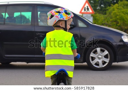 Little boy riding a bike on the road. Child is wearing reflective vest and helmet because of safety. Driving car in front of him. Sign of Children going to or from school in the background.