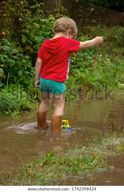 A
little boy in rubber boots pulls a toy toy car tractor on a rope
along a country sandy road with puddles after
rain.
