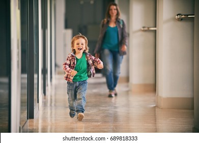 Little Boy Running In The Shopping Center With His Mom On Background.