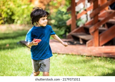 Little Boy Running With Toy Pistol In Yard Of Summer House On Summer Sunny Day Outdoors