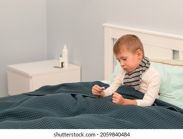 A little boy in a scarf is sick and lies in bed, holding a thermometer in his hands. Seasonal cold concept.  - Shutterstock ID 2059840484