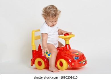 Little Boy Sit On Red Toy Car And Holding Tiny Vehicle In Hands, Looking On Floor, Barefoot Child Wearing Bodysuit Playing Isolated Over White Background.